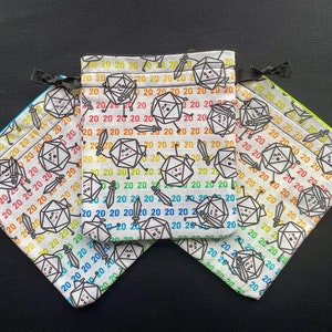 May include: Three white fabric dice bags with a repeating pattern of a cartoon dice with swords and the number 20. The bags are in different colours: green, yellow, and blue.
