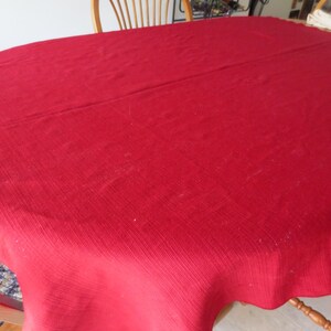 May include: A solid red tablecloth covers a wooden table. The tablecloth is a deep shade of red and has a subtle ribbed texture. The table is set with wooden chairs, suggesting a dining or kitchen setting.