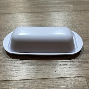 May include: A white rectangular butter dish with a lid. The dish has rounded edges and a smooth, glossy finish. The lid fits snugly on the base, creating a clean, minimalist design. The butter dish is designed to hold a standard stick of butter.