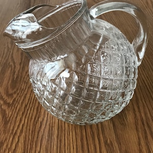 May include: A clear glass pitcher with a textured, diamond-patterned body and a curved handle. The pitcher has a spout for easy pouring. The pitcher is sitting on a wooden surface.