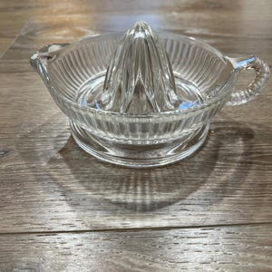 May include: A clear glass citrus juicer with a ribbed bowl, a conical reamer, and a small handle. The juicer sits on a round base. The kitchen tool is designed for squeezing fresh juice.