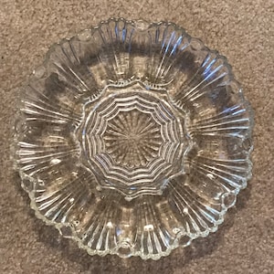 May include: A clear glass serving dish with a scalloped edge and a starburst design in the center. The dish has multiple compartments and a ridged pattern radiating from the center.
