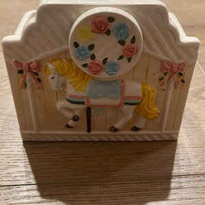 May include: A white ceramic carousel horse figurine with a pink, blue, and yellow floral wreath on the top. The horse is standing on a yellow base with a white picket fence design.