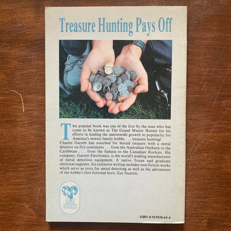 May include: A book cover with the title "Treasure Hunting Pays Off" and a photo of a person's hands holding a pile of coins. The book is by Charles Garrett, who is known as "The Grand Master Hunter" for his work in the treasure hunting hobby. The book cover also includes the text "RAM BOOKS" and the ISBN number 0-915920-65-4.