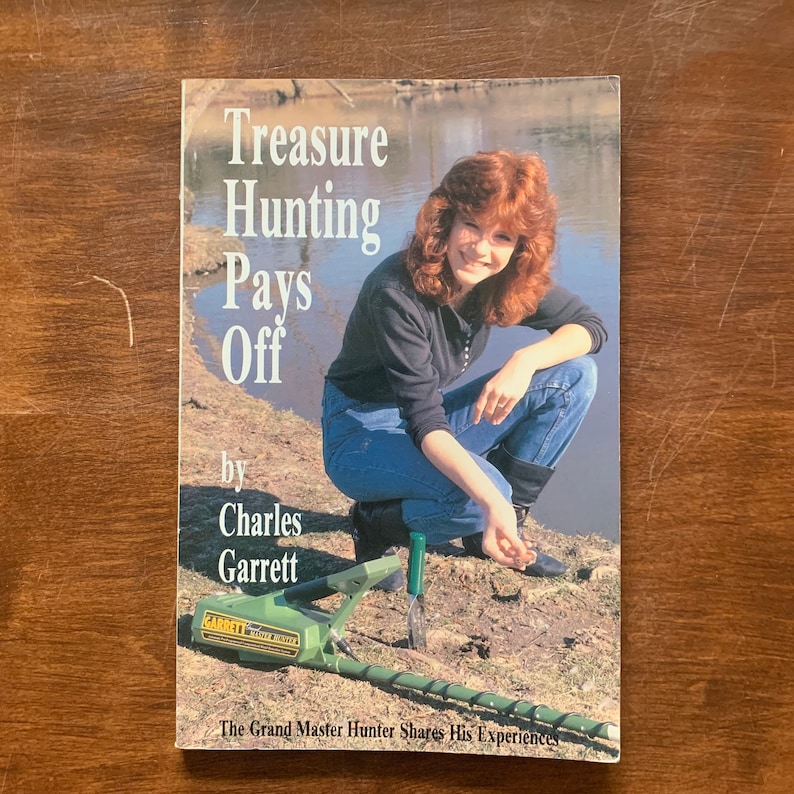 May include: A book titled "Treasure Hunting Pays Off" by Charles Garrett. The cover features a woman holding a metal detector and a small object in her hand. The book cover is green with white text.