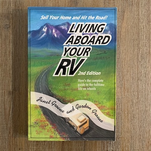 May include: Book cover with a scenic illustration of a winding road and mountains. The title is "Living Aboard Your RV" with the subtitle "2nd Edition". The authors are Janet Groene and Gordon Groene.