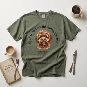 May include: An olive green t-shirt with a Cavapoo dog illustration and the text "excuse me while I snuggle my cavapoo". The image includes a coffee cup, a book titled "The Art of Slow Living", and cutlery.
