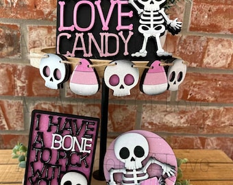 May include: A tiered Halloween-themed tray with decorative elements. It features a black sign with pink lettering that reads "Skeletons Love Candy," a skeleton figure, and a banner of skull and candy corn shapes. Additional signs say "I Have A Bone To Pick With You."