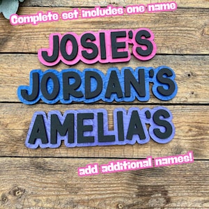 May include: Wooden name cutouts in pink, blue, and purple with black lettering. The names "JOSIE'S", "JORDAN'S", and "AMELIA'S" are displayed. Text at the top reads "Complete set includes one name" and at the bottom "add additional names!"