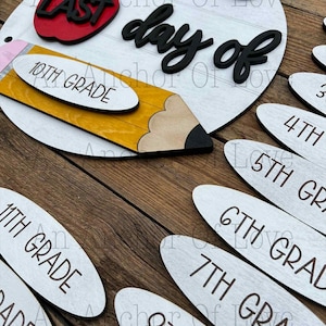 May include: A wooden school-themed sign with the text "Josie's Last Day of" and a pencil, apple, and grade level markers. The sign is white with black and red accents. Grade levels include 10th, 11th, 3rd, 4th, 5th, 6th, 7th, and 8th grade.