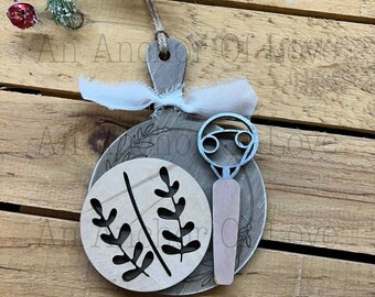 May include: Wooden Christmas ornament with a circular design. The ornament features a light brown color with a cut-out leaf design and a light pink bottle opener. A white ribbon and twine are attached for hanging. The text "An Anchor Of Love" is visible.