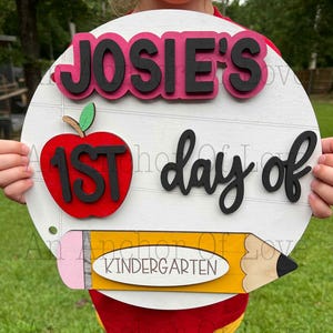 May include: A circular, white wooden sign with the words "JOSIE'S 1ST day of Kindergarten." The sign features a red apple with black lettering, a yellow pencil, and black text. The sign is held by a person.