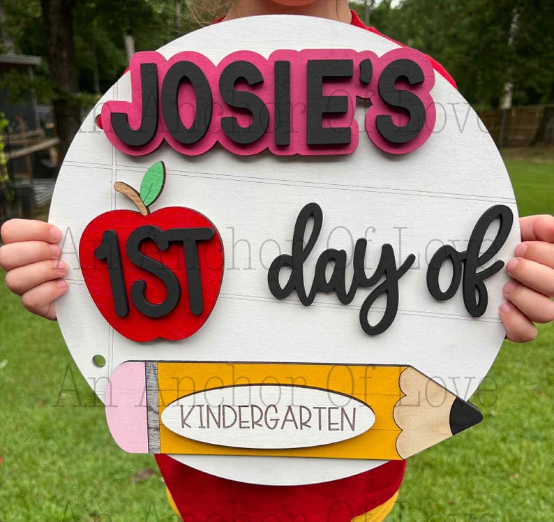 May include: A circular, white wooden sign with the words "JOSIE'S 1ST day of Kindergarten." The sign features a red apple with black lettering, a yellow pencil, and black text. The sign is held by a person.