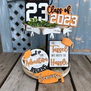 May include: A tiered tray decorated with graduation themed signs. The top tier features a black and orange sign that says "Senior 23" and a white sign that says "Class of 2023". The middle tier has three white signs with black text that say "Grad", "I'm Done", and "Finally". The bottom tier has a round white sign with black text that says "And so the adventure begins" and a black and white sign that says "The tassel was worth the hassle". There is also a small orange sign that says "Seniors".
