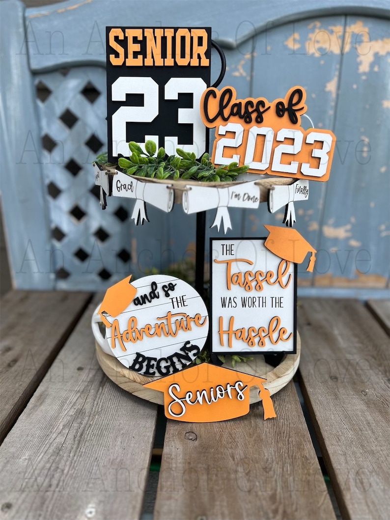 May include: A tiered tray decorated with graduation themed signs. The top tier features a black and orange sign that says "Senior 23" and a white sign that says "Class of 2023". The middle tier has three white signs with black text that say "Grad", "I'm Done", and "Finally". The bottom tier has a round white sign with black text that says "And so the adventure begins" and a black and white sign that says "The tassel was worth the hassle". There is also a small orange sign that says "Seniors".