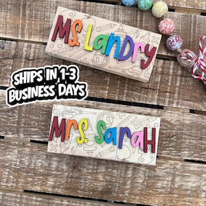 May include: Two wooden teacher name signs. The top sign reads "Ms. Landry" in colorful letters, with school-themed illustrations. The bottom sign reads "Mrs. Sarah" in colorful letters, with school-themed illustrations. The signs are on a wooden surface.