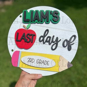 May include: A circular wooden sign with the words "LIAM'S LAST day of 3RD GRADE". The design includes a red apple, a yellow pencil, and green detailing. The background is white with horizontal lines.