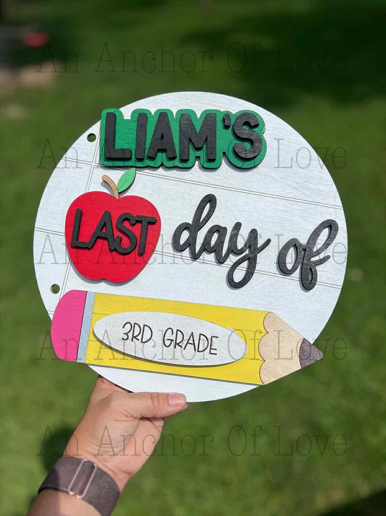 May include: A circular wooden sign with the words "LIAM'S LAST day of 3RD GRADE". The design includes a red apple, a yellow pencil, and green detailing. The background is white with horizontal lines.