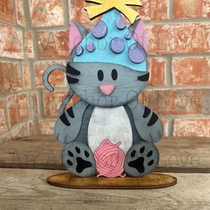 May include: A gray cat wearing a blue party hat with yellow stars and purple polka dots. The cat is holding a pink ball of yarn and has black paw prints on its feet. The cat is sitting on a wooden base.