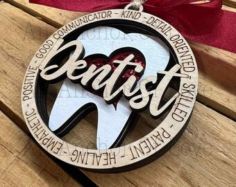 May include: A wooden dentist ornament with a red ribbon. The ornament features the word "Dentist" in a cursive font, a tooth graphic, and words like "Kind" and "Skilled" around the edge. The center has a heart filled with red glitter.