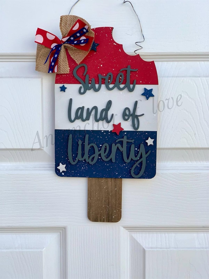 May include: A wooden popsicle-shaped door hanger with a red, white, and blue color scheme. It features the text "Sweet land of Liberty" with stars and a burlap bow.
