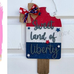 May include: A wooden popsicle-shaped sign with red, white, and blue paint and glitter. The sign says "Sweet land of Liberty" with stars and a brown stick at the bottom.