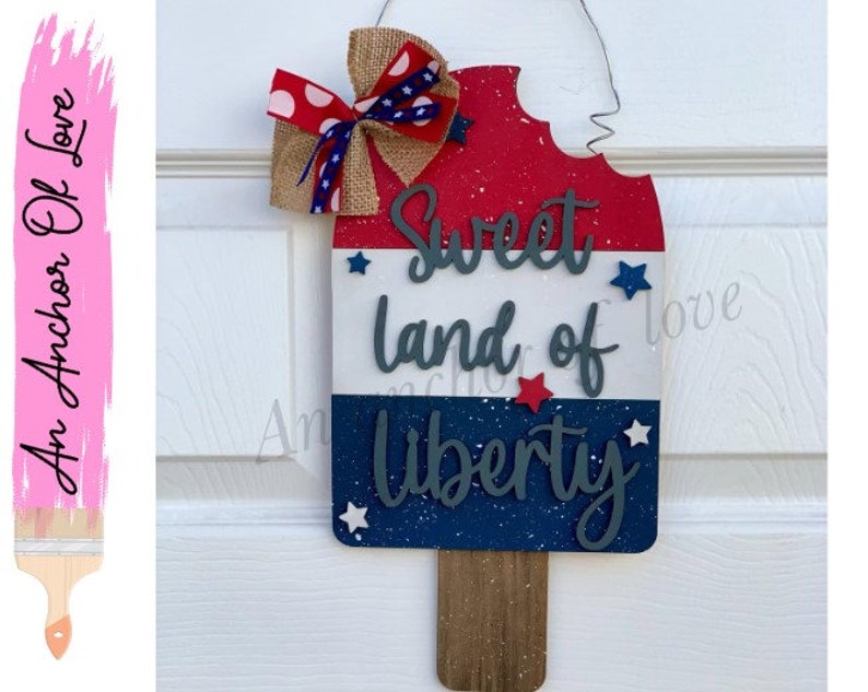 May include: A wooden popsicle-shaped sign with red, white, and blue paint and glitter. The sign says "Sweet land of Liberty" with stars and a brown stick at the bottom.