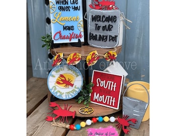 Pode incluir: Uma bandeja de três níveis decorada com um tema de camarão. O nível superior tem uma placa que diz "Quando a vida te dá limões, faça camarão". O nível intermediário tem uma placa que diz "Bem-vindo à nossa panela fervente" e uma placa que diz "Ponha um pouco de sul na sua boca". O nível inferior tem uma placa que diz "Não danço até lamber um pouco".