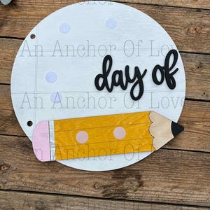 May include: A circular wooden sign with the words "day of" in black script. A yellow wooden pencil with a pink eraser and two pink dots is attached to the bottom. The sign has a notebook paper design with light blue dots.