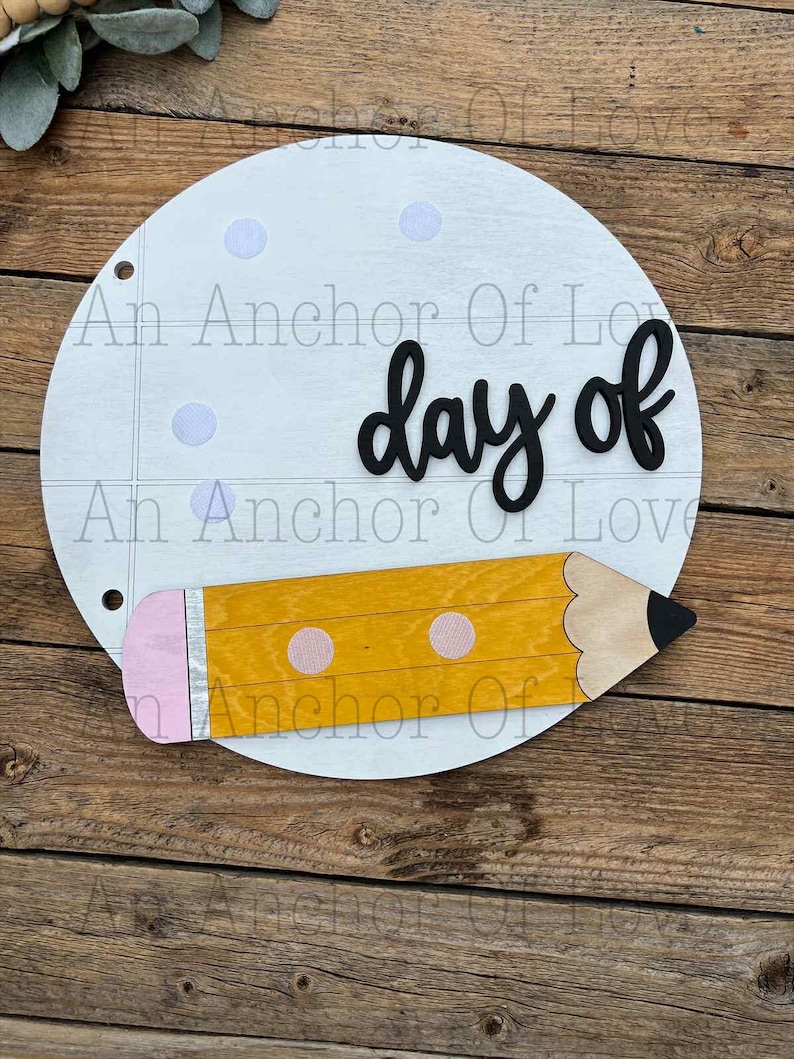 May include: A circular wooden sign with the words "day of" in black script. A yellow wooden pencil with a pink eraser and two pink dots is attached to the bottom. The sign has a notebook paper design with light blue dots.