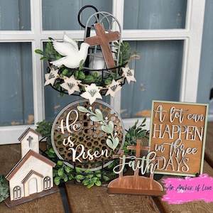 May include: A tiered wooden stand with a white dove, cross, and greenery. The top tier has a sign that says "He is Risen". The bottom tier has a sign that says "Faith". A wooden sign with the text "A lot can happen in three days" is on the right side of the stand.