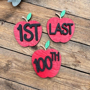 May include: Three wooden apple-shaped decorations. Each red apple has a black number or word: "1ST", "LAST", and "100TH". The apples have green leaves and brown stems. The background is a wooden surface.