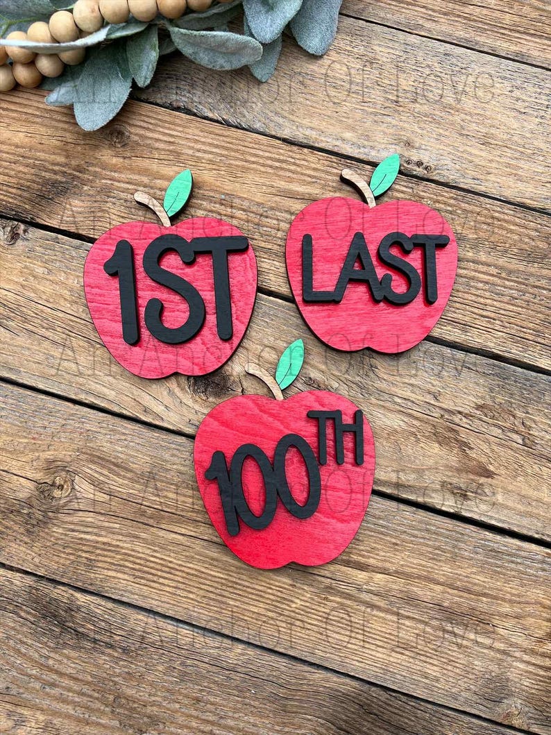May include: Three wooden apple-shaped decorations. Each red apple has a black number or word: "1ST", "LAST", and "100TH". The apples have green leaves and brown stems. The background is a wooden surface.