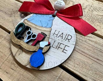 May include: Wooden Christmas ornament with a red bow and the text "HAIR LIFE." The ornament features a barber's tools, including scissors, combs, and a blue spray bottle. A wooden bead with "2025" is attached.