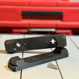 Może przedstawiać: Dwa czarne plastikowe uchwyty ścienne z otworami na śruby i mała sterta srebrnych śrub.