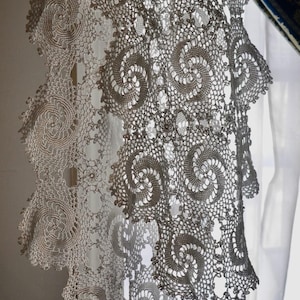 May include: Close-up of a delicate, off-white crocheted lace. The lace features a repeating spiral design with scalloped edges. The intricate pattern is visible, creating a textured and airy appearance. The lace is likely used for decorative purposes.