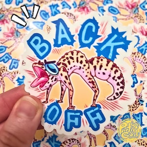 Può includere: Un adesivo con un geco dei cartoni animati con occhiali da sole e con la lingua di fuori. Il geco è marrone con macchie scure ed è arcuato. Le parole "BACK OFF" sono in lettere blu a bolle. Lo sfondo è pieno di adesivi simili.