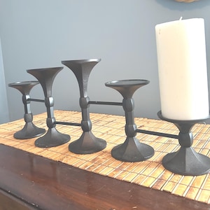 May include: A black metal candle holder with four connected stands. The stands have a flared top and a round base. A white pillar candle sits on the rightmost stand. The candle holder is on a bamboo mat.