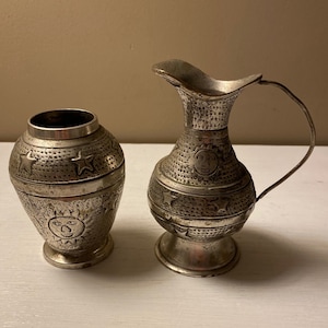 May include: Two silver metal decorative vases with embossed star and sun designs. The vase on the left is a cylindrical shape and the vase on the right is a pitcher with a handle.