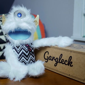 May include: A white, furry puppet with a single blue eye, gold ear accents, and a blue mouth. It wears a black and white zigzag patterned scarf. The puppet is next to a cardboard box with the word "Garglech" printed on it.