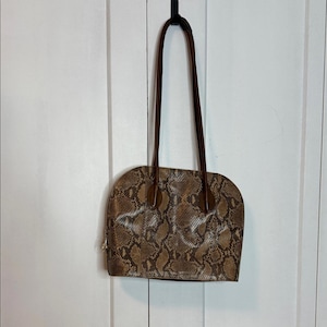 May include: A brown and tan faux snakeskin handbag with dark brown handles. The bag has a rounded top and a zip closure. The handles are long and thin, and the bag is hanging from a black hook.