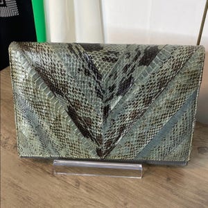 Peut inclure: Une pochette rectangulaire avec un motif en chevrons. La pochette est faite d'une matière semblable à de la peau de serpent dans des tons de vert, de gris et de marron. Le sac repose sur un présentoir en acrylique transparent.