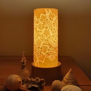 Puede incluir: Una lámpara cilíndrica con un diseño de encaje floral, iluminada desde dentro, que emite un brillo cálido. La lámpara se encuentra sobre una base de madera, con conchas decorativas y una pequeña botella de vidrio cerca. La lámpara mide unos 25 cm de alto.