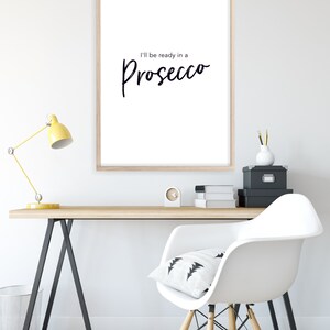 May include: A white framed print with the text "I'll be ready in a Prosecco" in black cursive font. The print is hanging on a white wall above a wooden desk with a yellow lamp, a white chair, and black storage boxes.