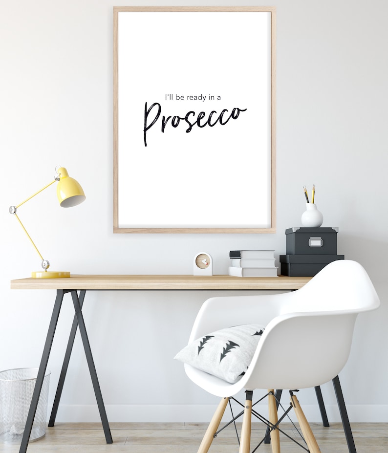 May include: A white framed print with the text "I'll be ready in a Prosecco" in black cursive font. The print is hanging on a white wall above a wooden desk with a yellow lamp, a white chair, and black storage boxes.