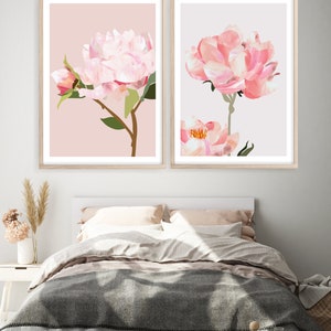 May include: Two framed prints of pink peonies on a light pink and light gray background. The prints are hanging on a white wall above a bed with a gray blanket.