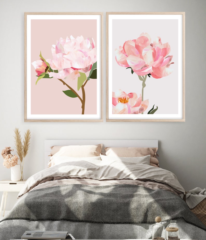 May include: Two framed prints of pink peonies on a light pink and light gray background. The prints are hanging on a white wall above a bed with a gray blanket.