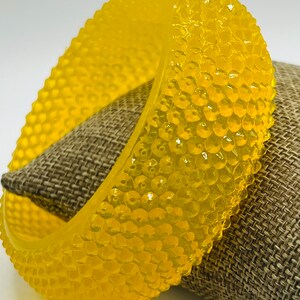 Op de afbeelding: Een felgele plastic armband met een textuur. De armband is gemaakt van een reeks kleine, verhoogde cirkels.