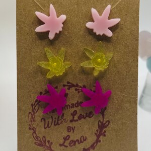 Puede incluir: Tres pares de pendientes con un diseño de hoja de marihuana. Los pendientes son de color rosa, amarillo y morado.