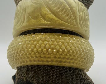 Könnte beinhalten: Zwei vintage gelbe Plastik-Armreifen. Der obere Armreif hat ein geschnitztes Blumenmuster. Der untere Armreif hat eine strukturierte Oberfläche.